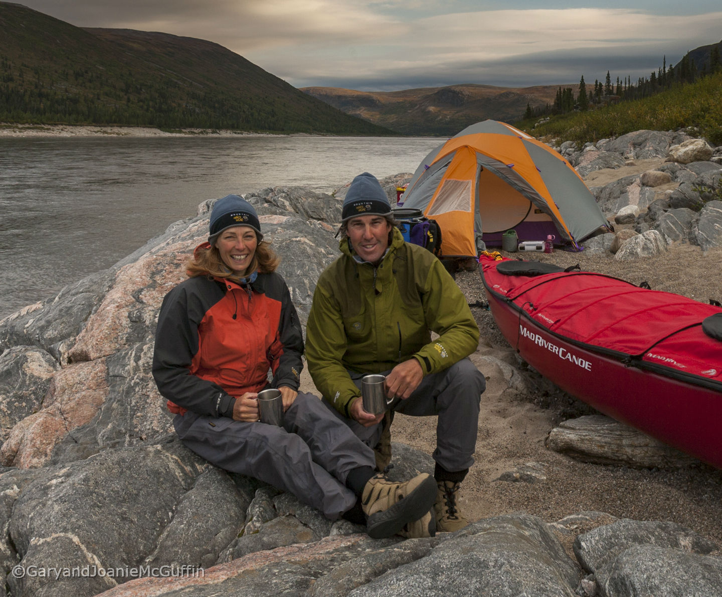 Couple sitting on rocks with their tent and canoe by a river
